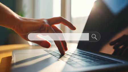 The a closeup of a hand typing on a laptop keyboard. A semi-transparent search bar with a magnifying glass is overlaid on the screen. The scene is brightly lit by sunlight streaming through a window, casting reflections and shadows on the laptop and workspace. The focus is on the hand and the laptop, suggesting a moment of digital search or work.の素材