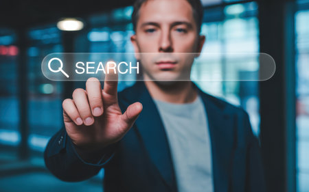 A man in a dark suit and jacket is touching a virtual search bar interface with his finger. The background is a blurred modern office setting with lights and glass, creating a futuristic and atmosphere. The man appears focused and serious, interacting with the digital search interface in an urban, corporate environment.の素材