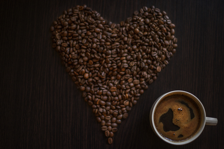 Coffee cup full of coffee and roasted coffee beans on wooden table  in the form of hearth. Coffee beans close up. Coffee background. Coffee addiction.の写真素材