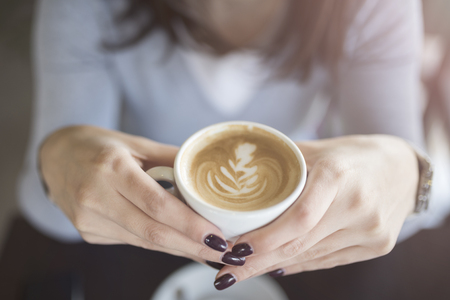 Woman`s hands holding cup of coffee with cream and foam on the brown wooden table in coffee shop.Top view;の写真素材