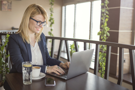 Business woman is working on computer  and drinking cup of coffee and glass of water in a coffee shop, restaurant. Charming happy woman student using laptop computer to prepare for the course work. Woman taking a coffee break, looking for information on internet.の写真素材