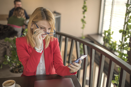 Young smiling blonde woman using smart phone, texting message in coffee shop, restaurant. Beautiful blonde with glasses using a smart phone and chatting with someone while drinking a cup of coffee.の写真素材