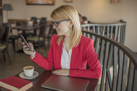 Young smiling blonde woman using smart phone, texting message in coffee shop, restaurant. Beautiful blonde with glasses using a smart phone and chatting with someone while drinking a cup of coffee.の写真素材