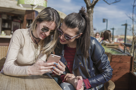 Two beautiful young women having fun outdoors while using their smartphones, friendship, happiness and people concept - two smiling girls whispering gossipの写真素材