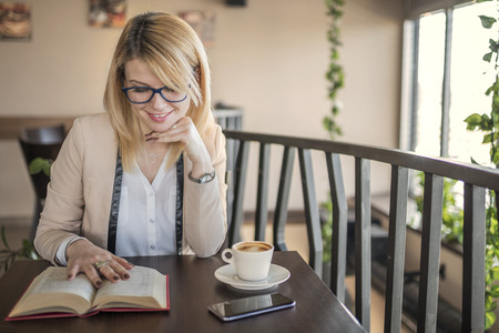Young smiling blonde woman with glasses in a restaurant reading a book and drinking coffee during a work break. Coffee break. Relaxing time. Business woman reading a book in coffee shop. Enjoy relax times with reading books caucasian woman.の写真素材