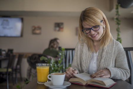 Young smiling blonde woman with glasses in a restaurant reading a book and drinking coffee during a work break. Coffee break. Relaxing time.  Enjoy relax times with reading book caucasian woman.の写真素材