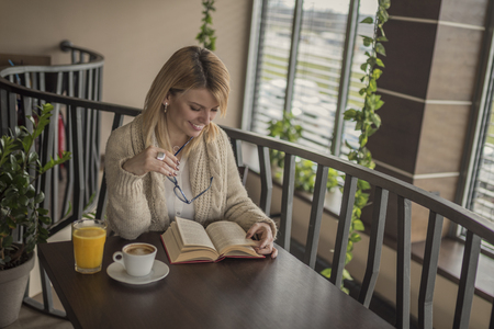 Young smiling blonde woman with glasses in a restaurant reading a book and drinking coffee during a work break. Coffee break. Relaxing time.  Enjoy relax times with reading book caucasian woman.の写真素材