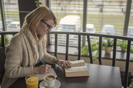 Young smiling blonde woman with glasses in a restaurant reading a book and taking notes during a work break. Coffee break. Relaxing time. Casual woman reading a book in coffee shop. Enjoy relax times with reading books caucasian woman.の写真素材