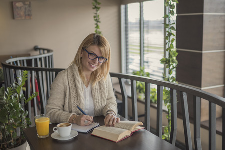 Young smiling blonde woman with glasses in a restaurant reading a book and taking notes during a work break. Coffee break. Relaxing time. Casual woman reading a book in coffee shop. Enjoy relax times with reading books caucasian woman.の写真素材