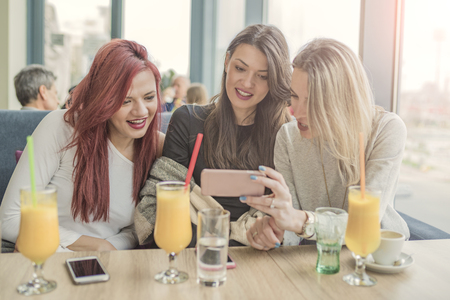 Portrait of three young beautiful women using mobile phone at coffee shop. Three young female friends enjoying together in a restaurant laughing and joking while watching funny videos on mobile phone.の写真素材