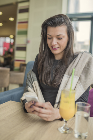 Young smiling brunette woman using smart phone, texting message in coffee shop, restaurant. Beautiful brunette using a smartphone and chatting with someone while drinking a cup of coffee.の写真素材