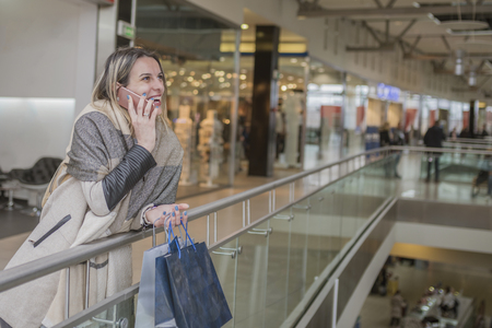Smiling girl talking on her mobile phone in shopping mall while holding shopping bags. Fashion Shopping Girl Portrait. Beauty Woman with Shopping Bags in Shopping Mall. Shopper. Sales. Shopping Centerの写真素材