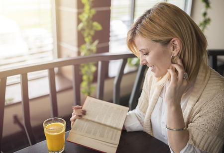 Young smiling blonde woman with glasses in a restaurant reading a book and drinking juice. Relaxing time.  Enjoying relax time and reading book .の写真素材