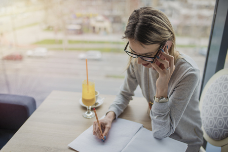 Business woman talking on smartphone, taking notes in a coffee shop, restaurant. Gorgeous woman taking a notes to prepare for the course work. Drinking juice at coffee shop.の写真素材