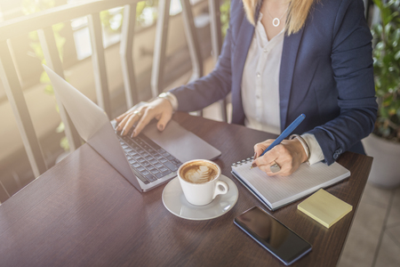 Business woman taking notes and using laptop in restaurant. Blonde girl solving business problems in coffee shop. Woman in blue suit.の写真素材