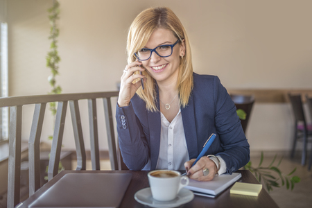 Attractive woman with glasses talking on smartphone, taking notes in a coffee shop, restaurant. Charming happy girl is preparing for work. Looking for information. Drinking coffee at coffee shop.の写真素材