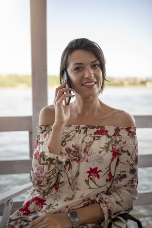 Portrait of beautiful young girl with phone on sunny day in nature, near river. Cheerful woman model sitting in cafe enjoying free time. Pretty girl talking on mobile phone.の写真素材