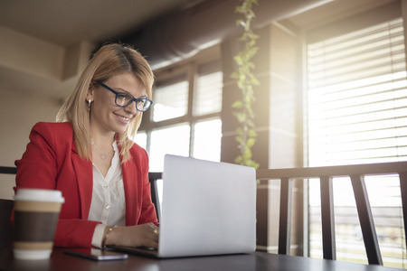 Business woman is working on laptop outdoor, in restaurant and drinking coffee to go. Charming happy girl is using laptop computer to prepare for work. Pretty girl with glasses is looking for information on internet.の写真素材