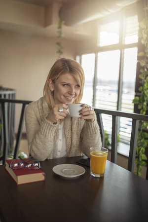 Young smiling blonde woman with glasses in a restaurant reading a book and drinking coffee. Relaxing time.  Enjoying relax time and reading book.の写真素材