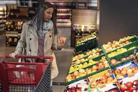 Housewives, women with shopping cart in supermarket. Gorgeous young woman with a shopping cart looking at some products on a supermarket aisle. Woman in the supermarket. Beautiful young woman shopping in a supermarket and buying fresh organic vegetables.の写真素材