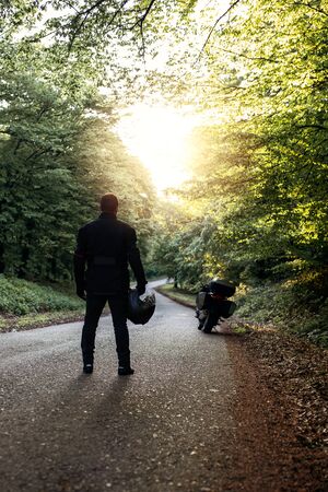 Motorcycle guy in raiders clothes, gearing up. A young men stands near an motorcycle in nature during sunset. Biker with motorcycle on the road. Motorcycle and safe driving. Helmet and motorcycle.の写真素材