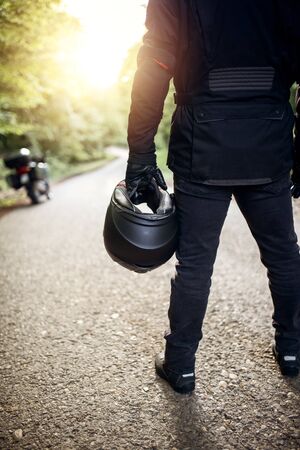 Motorcycle guy in raiders clothes, gearing up. A young men stands near an motorcycle in nature during sunset. Biker with motorcycle on the road. Motorcycle and safe driving. Helmet and motorcycle.の写真素材
