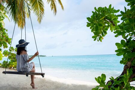 Young beautiful woman relaxing in swing hanging on coconut palm at exotic beach. Blue sea and white sand. Fashion girl enjoying on seashore. Portrait of young brunette relaxing on the beach.の写真素材