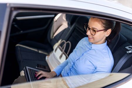 Attractive businesswoman with laptop in the back seat of the car. Close up portrait of a young business woman using laptop.の写真素材