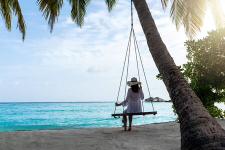 Young beautiful woman relaxing in swing hanging on coconut palm at exotic beach.Blue sea and white sand. Fashion girl enjoying on seashore. Brunette girl in beach dress on the beach, by the ocean.の写真素材