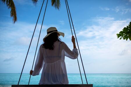 Young beautiful woman relaxing in swing hanging on coconut palm at exotic beach.Blue sea and white sand. Fashion girl enjoying on seashore. Brunette girl in beach dress on the beach, by the ocean.の写真素材