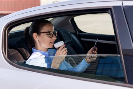 Attractive young woman on the business trip, taking a coffee break. Attractive business woman with phone in the back seat of the car. Close up portrait of a young business woman.の写真素材