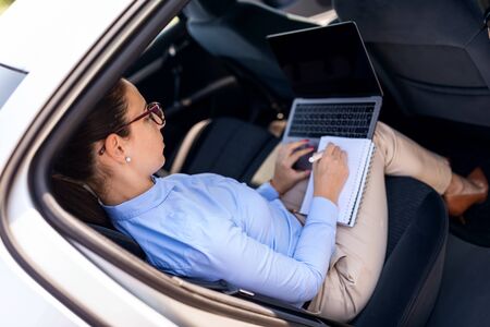 Attractive businesswoman with laptop in the back seat of the car. Close up portrait of a young business woman using laptop and taking notes.の写真素材