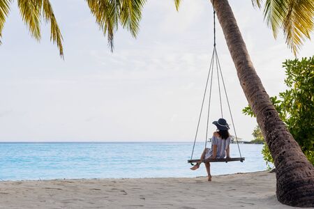 Beautiful fit girl on travel vacation. Portrait of smiling and attractive young woman at sea. Young beautiful woman relaxing in swing hanging on coconut palm at exotic beach.の写真素材