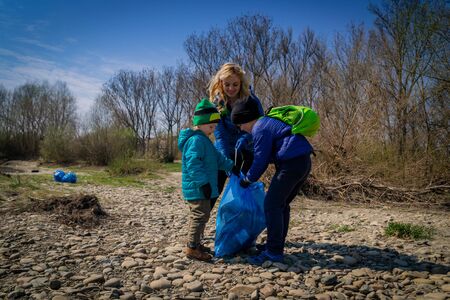 young blond hair woman with her sons in blue gloves cleaning the river bank from plastic and garbage in big blue trash bags on a sunny day in early springの写真素材