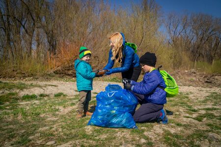 young blond hair woman with her sons in blue gloves cleaning the river bank from plastic and garbage in big blue trash bags on a sunny day in early springの写真素材