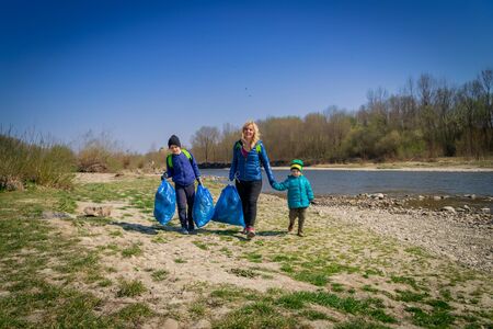 young blond hair woman with her sons in blue gloves cleaning the river bank from plastic and garbage in big blue trash bags on a sunny day in early springの写真素材