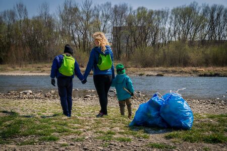 young blond hair woman with her sons in blue gloves cleaning the river bank from plastic and garbage in big blue trash bags on a sunny day in early springの写真素材