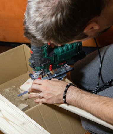man using electric jigsaw at home to cut piece of wood board on a cardboard box. Do it yourself.の写真素材