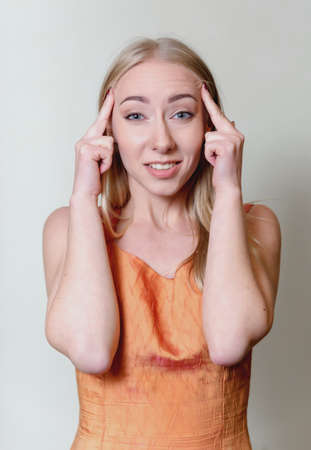 beautiful young blonde woman holding fingers on pulse trying to think looking at camera woman in orange sleeveless dress photo on light backgroundの写真素材