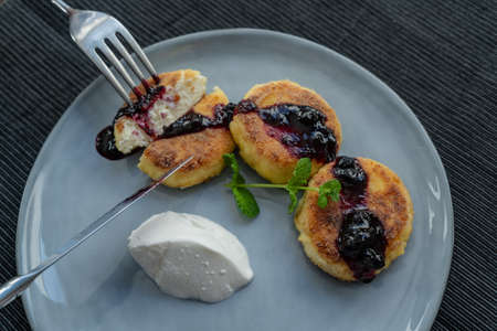 Delicious fried cheesecakes with a crispy crust, poured with cherry jam, served on a gray ceramic plate with a ball of sour cream and decorated with a sprig of fragrant mintの写真素材