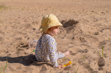 little girl in a yellow panama sitting on the sand on the beach near the water in sunny summer weather the child is enjoying the rest playing with the sandの写真素材