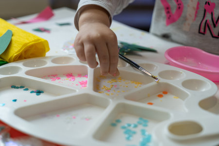 a child's hand sorts shiny colored pieces for decoration in a white paint paletteの写真素材