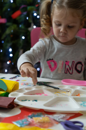 A little girl makes and decorates Christmas tree decorations with colored paper and sparklesの写真素材