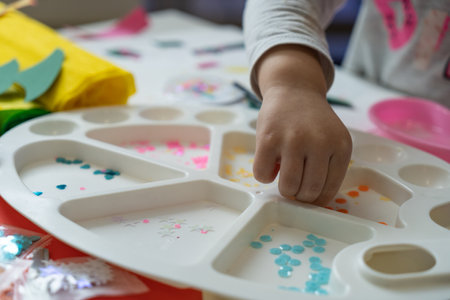 a child's hand sorts shiny colored pieces for decoration in a white paint paletteの写真素材