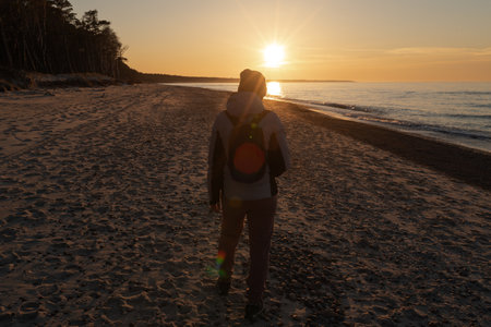 Back view of young woman with backpack standing on the beach at sunsetの写真素材