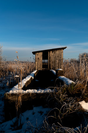 Old wooden water well and abandoned house in the winter field with snow and blue skyの写真素材