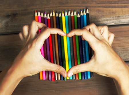Female hands in the shape of a heart on colored pencils on a background of dark wood tables