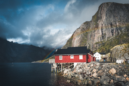 Rainbow ofer red houses rorbuer of Reine in Lofoten, Norway with red rorbu houses, clouds, rainy blue sky and sunny. Bridges and mountains in the background.の写真素材