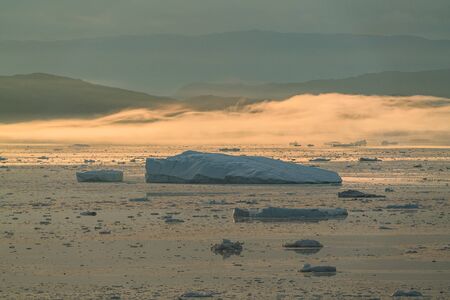 Stranded icebergs in the fog at the mouth of the Icefjord near Ilulissat. Nature and landscapes of Greenland. Travel on the vessel among ices. Phenomenon of global warming. Coast in the sunset.の写真素材