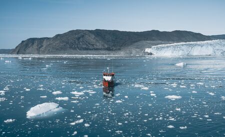 Red Passenger cruise ship sailing through the icy waters of Qasigiannguit, Greenland with Eqip Sermia Eqi Glacier in Background. A small boat among icebergs. Sailboat cruising among floating icebergs in Disko Bay glacier during midnight sun Ilulissat, Greenland. Studying of a phenomenon of global warming Ices and icebergsの写真素材